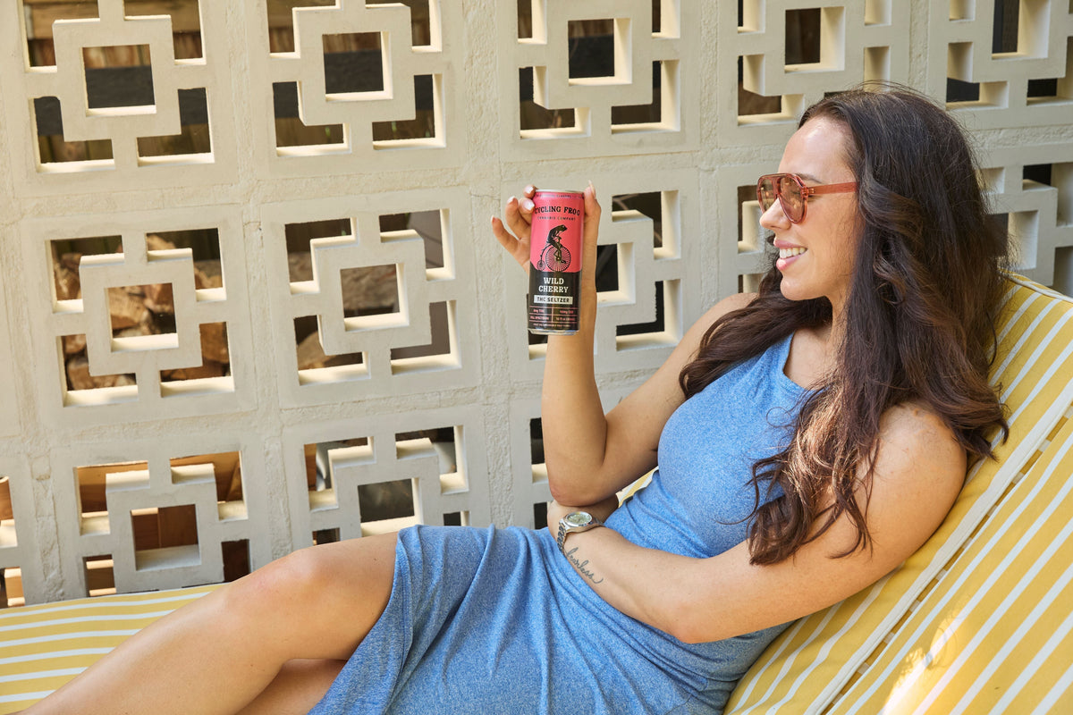 woman in red sunglasses and a blue dress relaxing on a yellow and white striped lawn chair holding up a Cycling Frog Wild Cherry THC Seltzer and smiling.