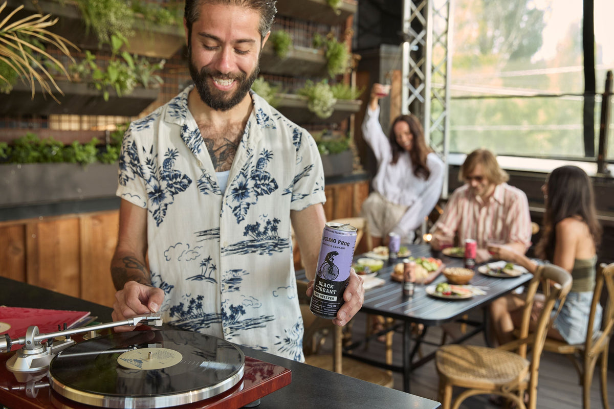 a person spinning a vinyl record on a turntable and holding a can of Black Currant Cycling Frog 5mg THC  + 10mg cbd, while a table of people dine, converse, and drink cycling frog in the background.